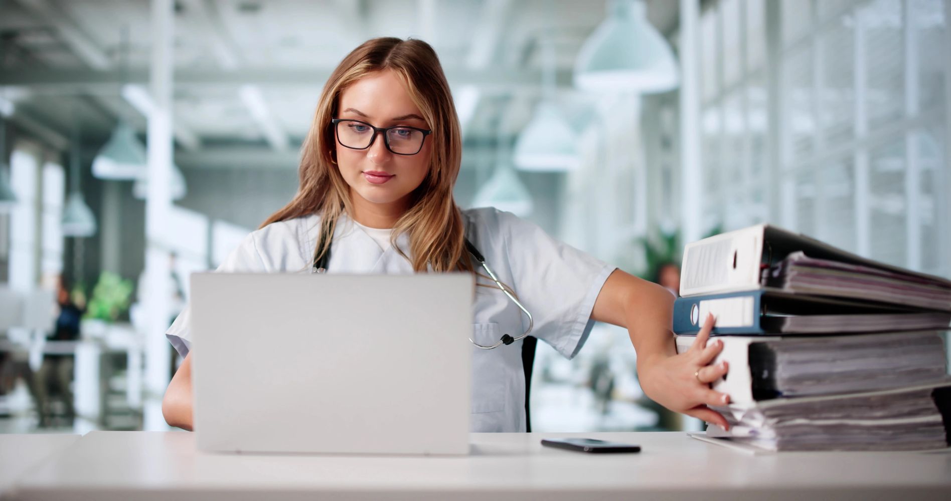 Medical professional working on a laptop with a stack of files beside her.