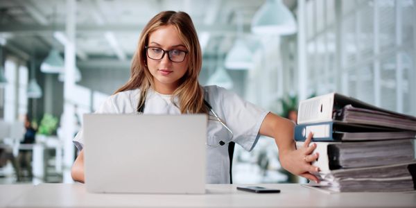 Medical professional working on a laptop with a stack of files beside her.