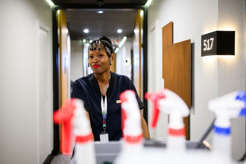 Confident young woman with braided hair pushing cleaning trolley down hallway, with vibrant spray bottles in foreground and room number signage visible in background.