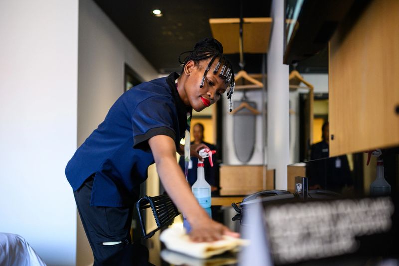 Side view of focused cleaner wiping a glossy table surface with a yellow cloth in a hotel guest room, cleaning products and hanging rails visible in softly lit setting.