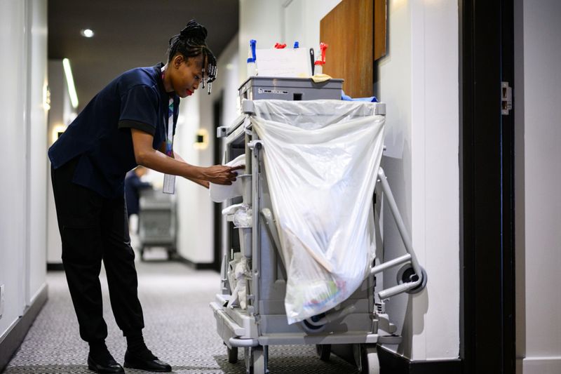 Side view of female hotel cleaner arranging linen items on a housekeeping trolley while working in a quiet hotel hallway lined with rooms and neutral decor.