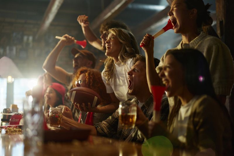 Large group of cheerful rugby fans having fun while watching a successful match on TV in a pub. Focus is on man with ball.