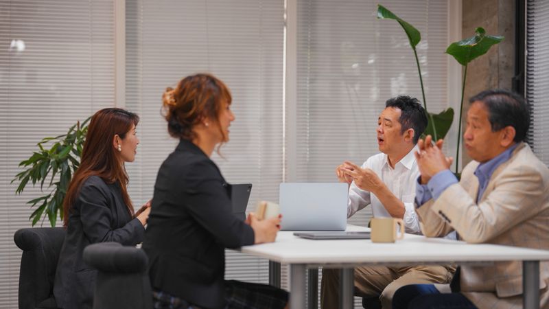 Four diverse office workers are sitting around a table and having a business meeting with their laptops and digital tablets in an office. A deaf businessman is using sign language to speak. There is a non-binary business person on the team.
