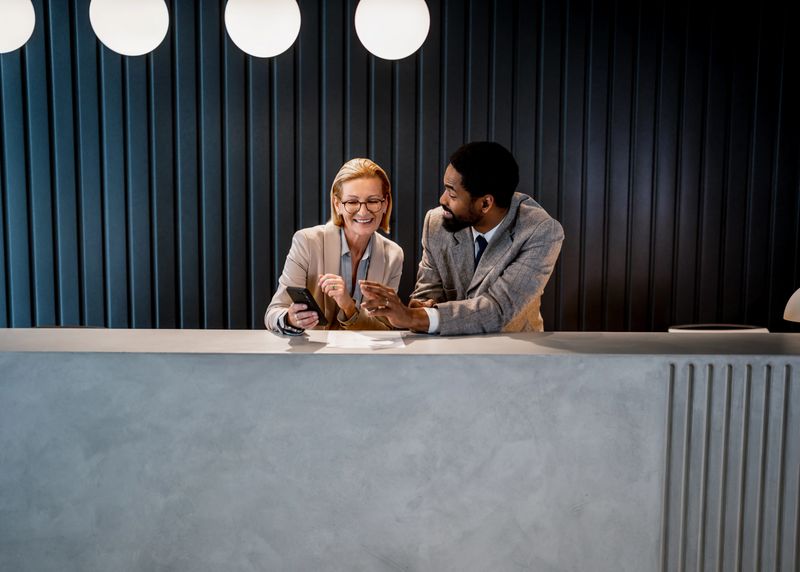 Two smiling hotel receptionists engaging with a smartphone while providing excellent customer service at the front desk in a modern lobby