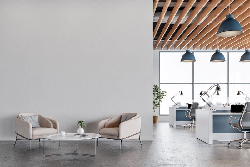 Stylish and contemporary office space featuring a cozy lounge corner with beige armchairs and a coffee table, set against a white smoke wall on cement floor. The open-plan workspace includes desks, office chairs, and task lighting under a ceiling with exposed wooden slats. Large windows bring in natural light, creating a bright and professional environment ideal for business or coworking use. 3D rendered image.