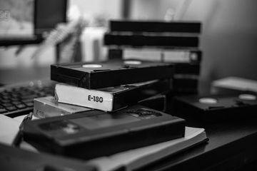 Stack of vintage VHS tapes on a desk with a keyboard and plant in the background.