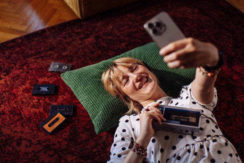 A cheerful woman, lying on a red rug with a green pillow, enjoys listening to a cassette tape on a vintage cassette player at home and taking selfies