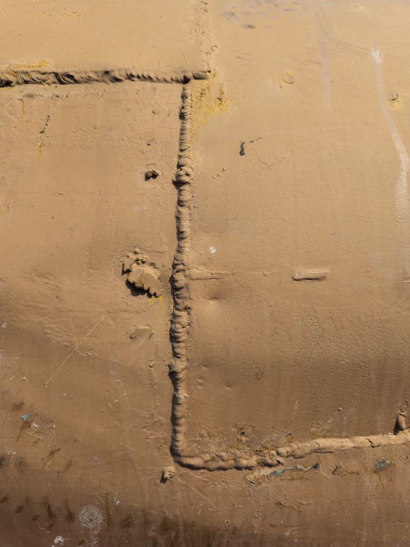 Rough, weathered metal surface showcases intricate welds under bright daylight. Industrial atmosphere highlights craftsmanship and outdoor elements with no people present.