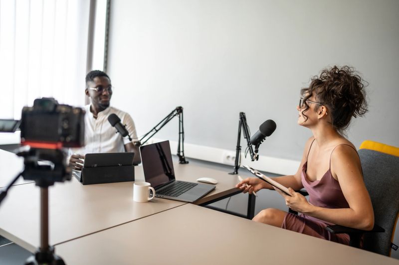 Two individuals engage in recording a podcast conversation in a modern studio with microphones and cameras, illustrating professionalism, creativity, and collaboration within a technologically advanced workspace.