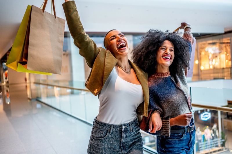 Two happy female customers are raising their shopping bags and laughing, enjoying their successful shopping spree in a mall