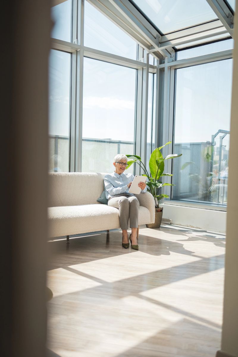 An older woman sits attentively on a couch in a spacious, sunlit conservatory, reading documents. The modern and clean living space creates a calm and peaceful environment, perfect for relaxation and focus.