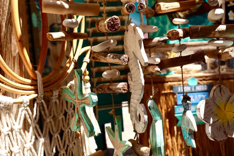 Handmade beach-themed wooden decorations, including a seahorse, crosses, and tropical elements, hang in bright sunlight at a coastal craft market.