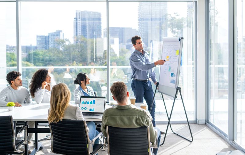 Business people watching a presentation on the whiteboard. A man is writing on the whiteboard with charts and graphs. They are sitting in a board room, there are laptop computers and technology on the table. Finance charts and graphs can be seen. There is a window behind him with city views.