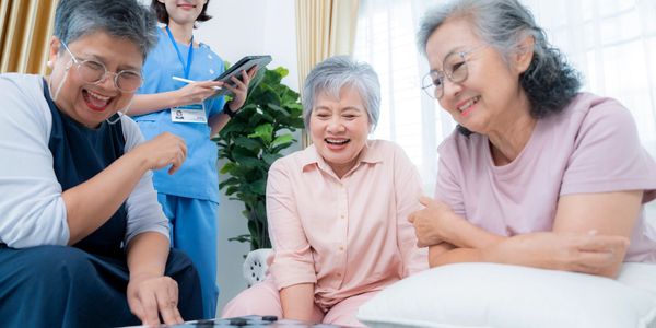 Three elderly women joyfully playing checkers with a nurse observing.