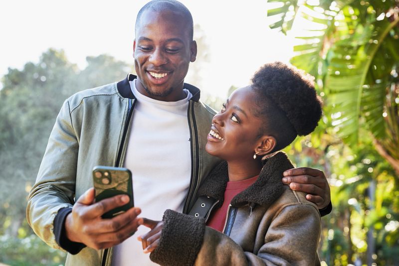 A joyful couple is seen enjoying a sunny day in a city park, embracing each other as they look at a smartphone. The lush greenery around them highlights their happiness and connection, symbolizing love and togetherness. The scene captures a candid and warm moment, perfect for lifestyle and relationship themes.
