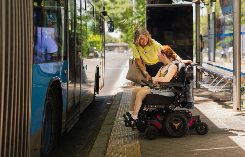 Smiling caregiver assisting a young red haired woman in a wheelchair as she boards a bus at a city stop, engaging in cheerful conversation and sharing moments of joy together