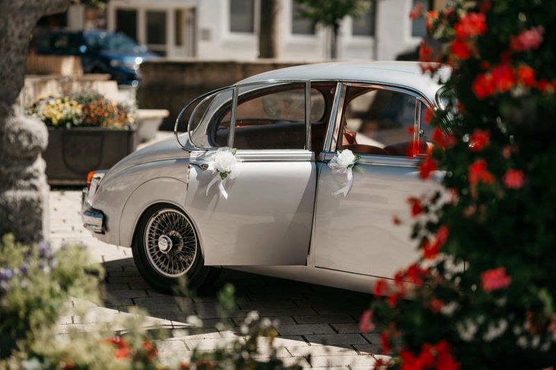 Classic vintage car with wedding decorations parked in a sunny courtyard, surrounded by vibrant flowers.