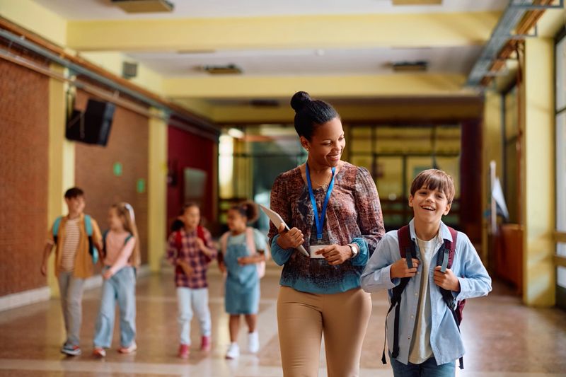 Happy boy and African American teacher talking while walking through hallway at school.