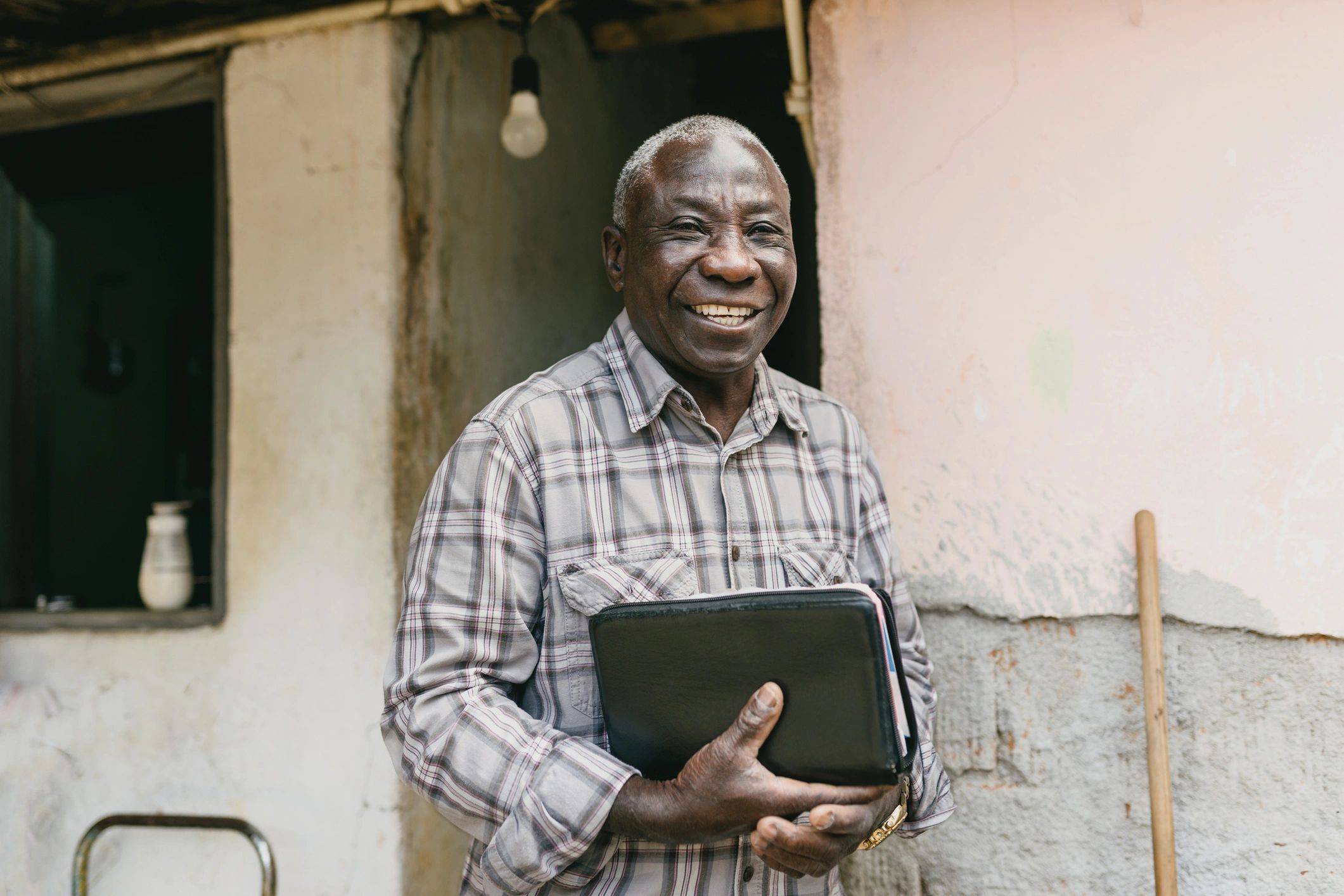 Smiling elderly man with a folder standing outside a rustic building.