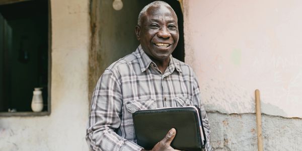 Smiling elderly man holding a book, standing by a rustic wall.