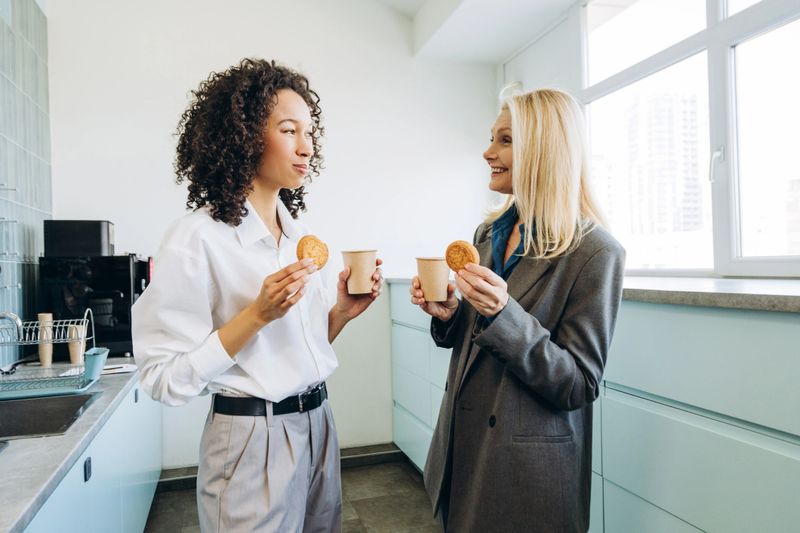 Two female colleagues enjoying coffee break in the office kitchen, smiling and chatting while holding cookies and paper cups, creating a warm and friendly atmosphere. Relaxing, business concept