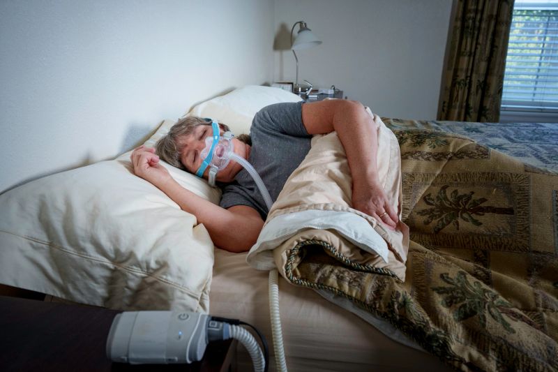 A person sleeps in bed while wearing a CPAP mask for sleep apnea treatment. The bedroom is dimly lit with a bedside lamp visible in the background. The CPAP machine sits beside the bed with tubing connected to the facial mask. The person is covered with a patterned bedspread and white pillows in a home setting. The window blinds are closed, creating a private sleeping environment.