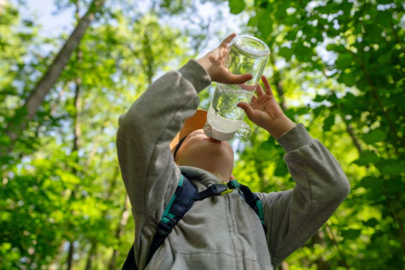 A child wearing outdoor attire drinks water from a bottle while enjoying a forest environment, emphasizing hydration, nature, and adventure.
