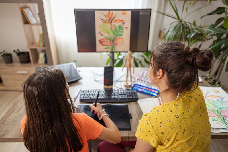 From an over-the-shoulder perspective, an art teacher points to a digital botanical drawing on a monitor, guiding her student who is actively working on a graphics tablet. Instruction. Computer. Monitor. Drawing. Skills. Collaboration. Home Studio. Over-the-Shoulder. Perspective. Botanical. Plants. Expression. Technology. Modern. Workspace.