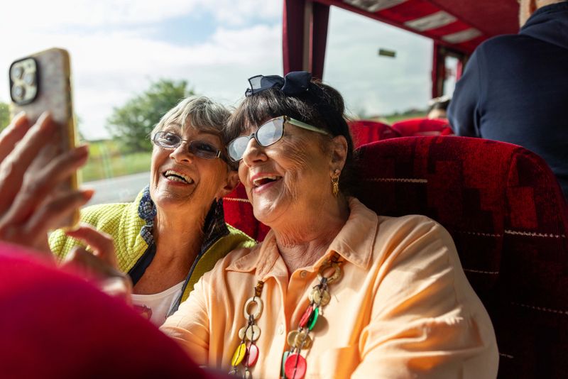 A close-up view of two senior female friends sitting side by side on a coach bus. They are wearing casual clothing. They have a cheerful expression, and they are smiling while taking a selfie on a smartphone.

Videos are available for this scenario.
