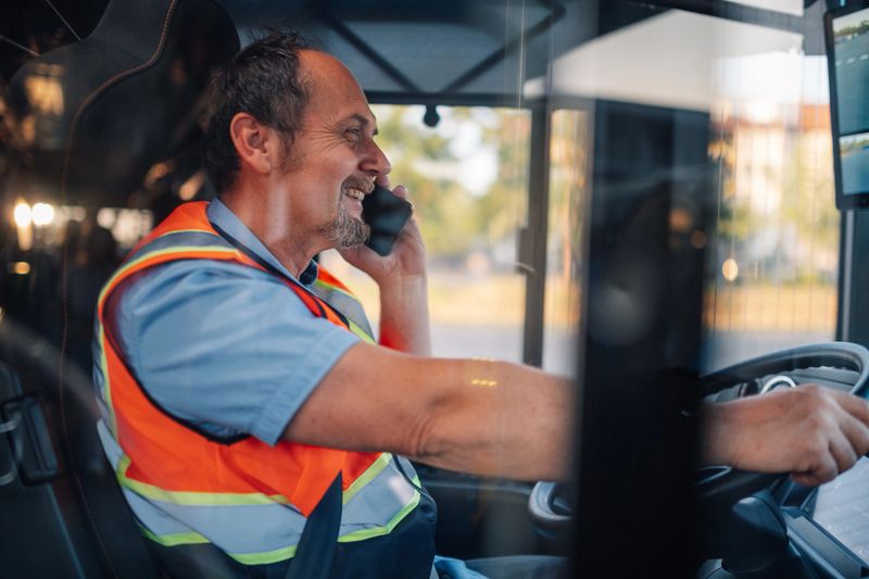 Smiling bus driver engaging in a conversation on a smartphone while adjusting the gps system, skillfully navigating the urban streets