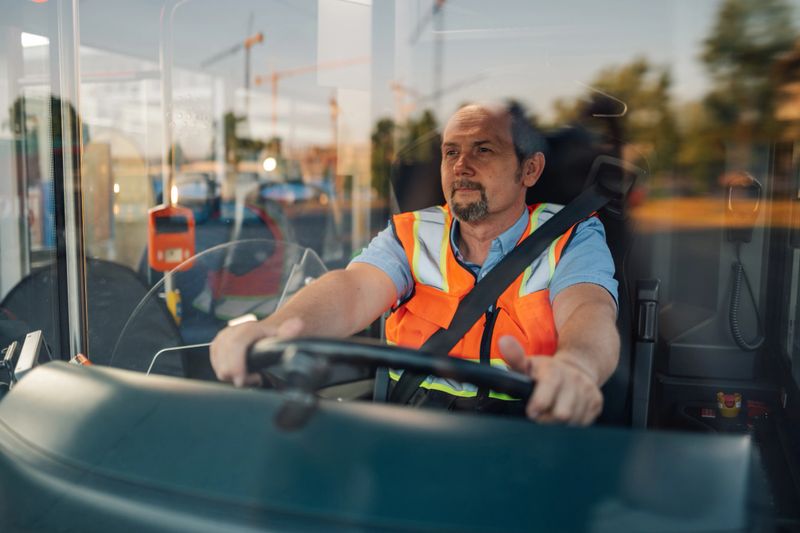 Mature bus driver wearing an orange safety vest and seatbelt, navigating the city streets with focus and responsibility while transporting passengers