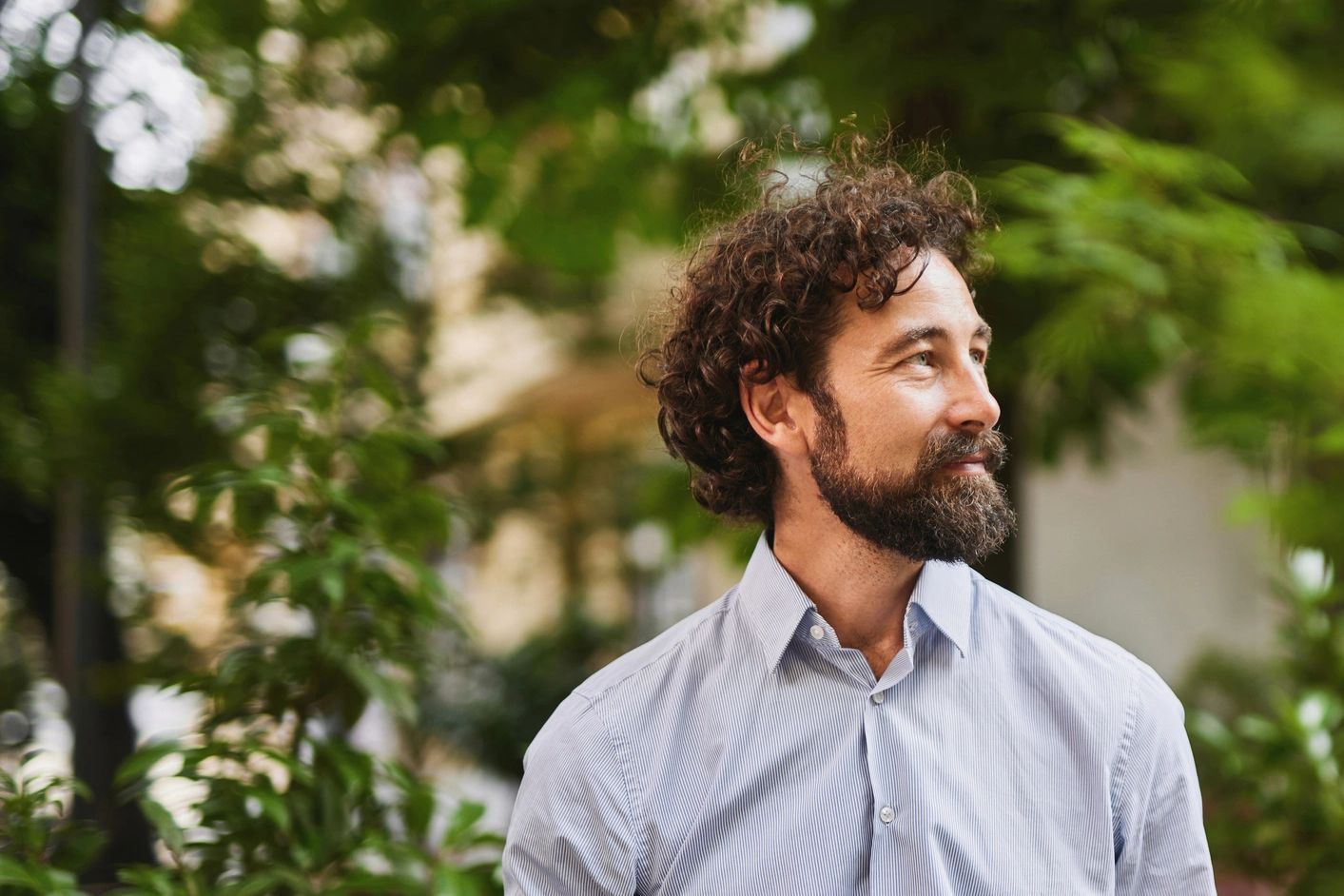Smiling bearded man outdoors surrounded by greenery.