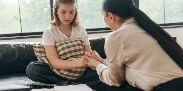A woman comforts a sad girl sitting on a couch, holding her hands.