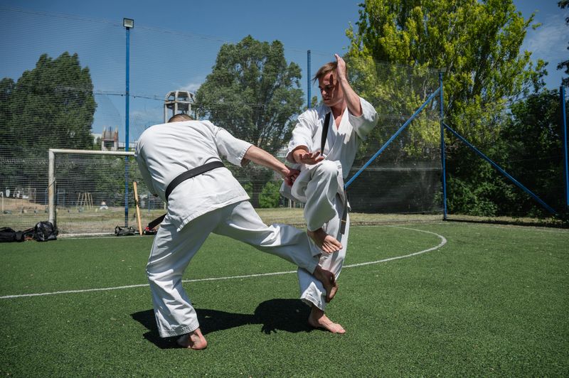 Man in white karate gi executing a leg sweep technique on his partner during martial arts training on a grassy field in sunny weather.