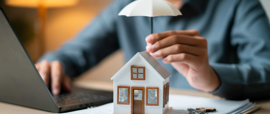 Person holding umbrella over model house near laptop.