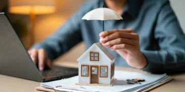Person holding an umbrella over a miniature house model on a desk.