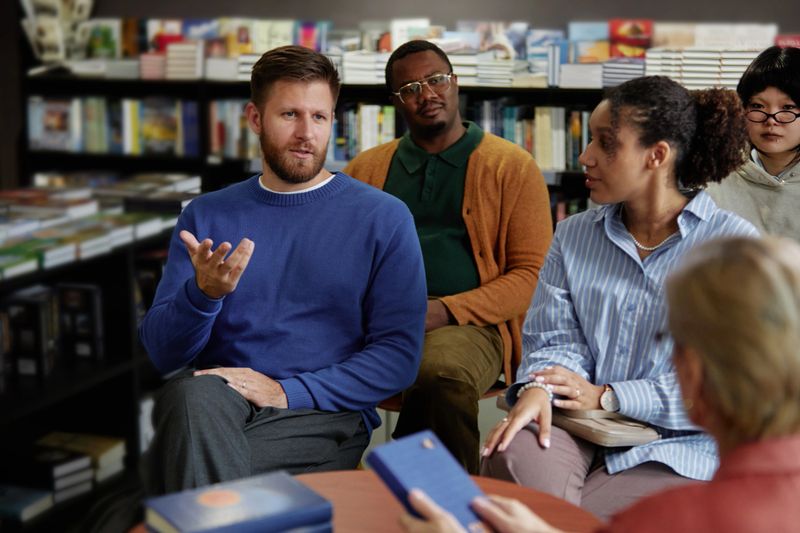 Caucasian middle aged man speaking during group discussion with Black middle aged man, Black young adult woman and Asian teenager listening in bookstore setting during session