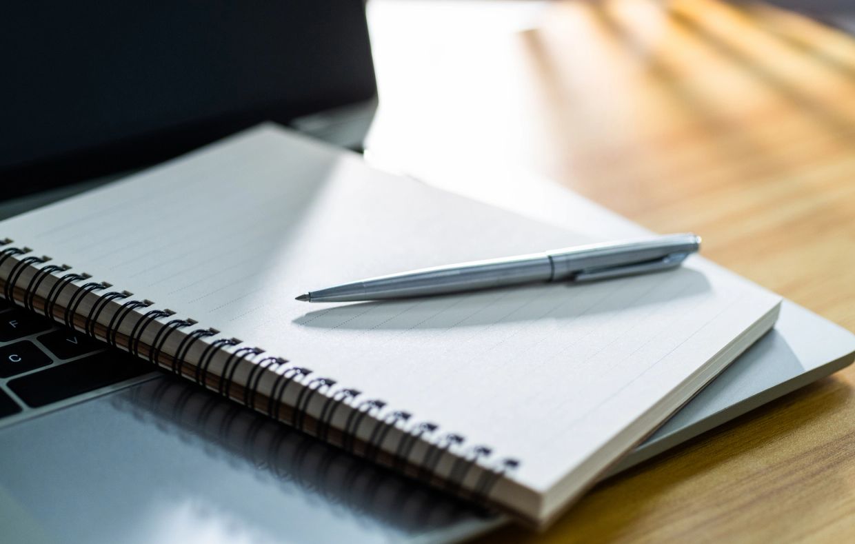 A silver pen rests on an open spiral notebook atop a laptop keyboard.