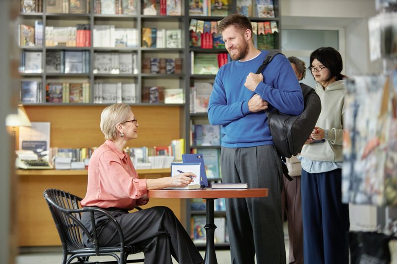 Middle aged Caucasian woman sitting at table signing book for man standing with backpack while Asian woman waiting in line inside bookstore