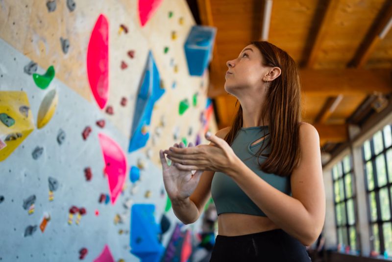 Young athletic woman applies climbing chalk to her hands in a moment of focus and preparation before attempting a route on an indoor bouldering wall. Brightly colored climbing holds fill the background of the modern gym, creating a vibrant and energetic setting for this motivational and fitness-themed scene.