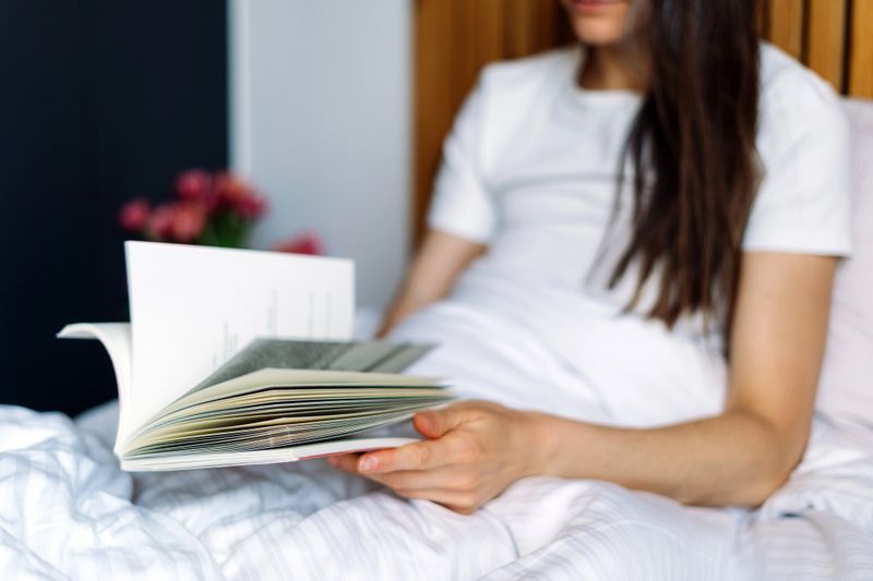 In a stylish hotel room, a guest relaxes on a cozy bed, flipping paper pages, reading a book. Woman surrounded by modern decor and soft linens, creating a tranquil atmosphere for stay