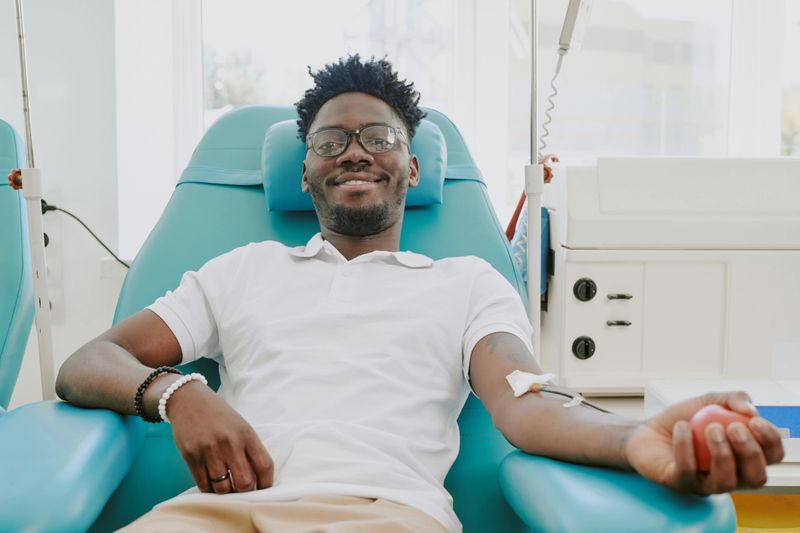 Young adult Black man donating blood while reclining in medical chair, smiling and looking relaxed, intravenous needle inserted in arm, participating in blood donation process at clinic
