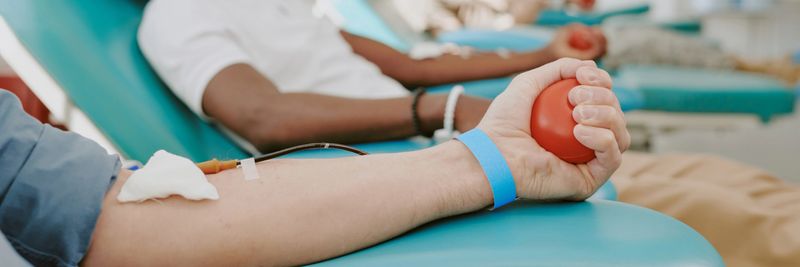 Caucasian man arm and Black man arm donating blood in medical facility, both lying on chairs with intravenous lines inserted, close up of hands holding stress balls during donation process