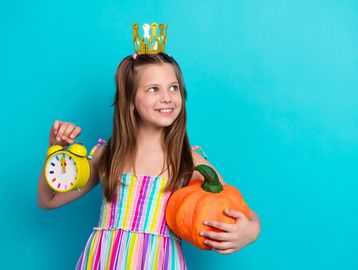 Girl wearing a crown holding a pumpkin and yellow alarm clock.