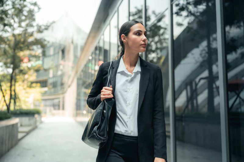 Cheerful young woman on her way to her first job interview pausing to check her appearance, adjusting her suit and smoothing her hair, glancing into a mirror window to ensure every detail is perfect, determined to make a confident and polished first impression and present the best version of herself