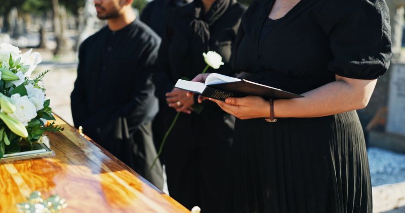 Bible, flower and woman by coffin at funeral for respect, blessing or spiritual comfort at graveyard. Religion, reading and person by casket with white rose and holy book for grief, loss or farewell.