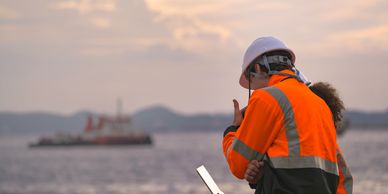 Workers in safety gear using a laptop near the water at sunset.