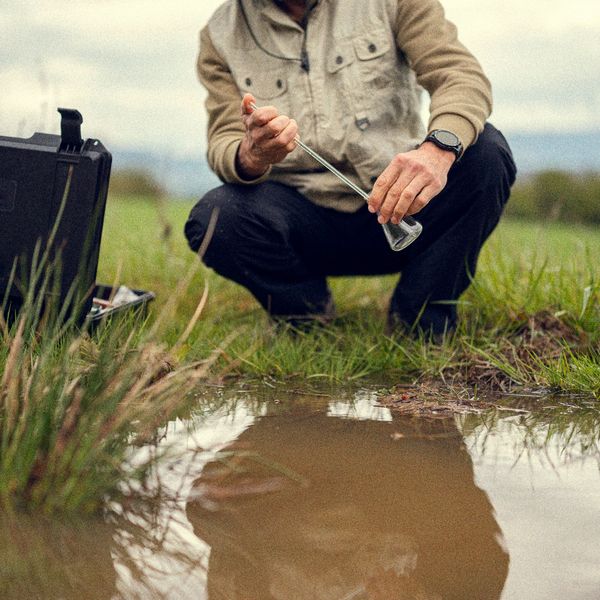 Person collecting water sample from a puddle outdoors.