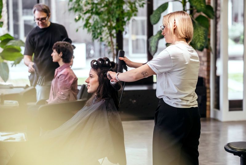 Side-view portrait of young female hairstylist in white shirt and black pants styling client's dark hair with professional tools in bright, modern salon environment.