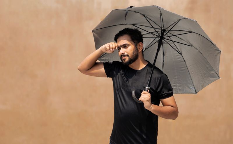 A man in a black Nike t-shirt stands outdoors on a sunny day, shielding himself from the sun with a dark gray umbrella.  The background is a muted earth-toned wall.